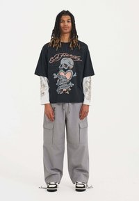 Schwarz übergroßes T-Shirt mit einem bunten Totenkopf- und Herz-Graphic; kombiniert mit lockeren grauen Cargo-Hosen und schwarz-weißen Sneakers.