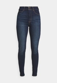 Jeans de mezclilla azul oscuro de talle alto y ajuste slim, con dos bolsillos frontales, costuras amarillas y cierre de botón.