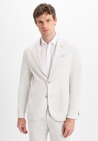 Light gray linen blazer with two front pockets, notched lapels, and two buttons. Worn over a white shirt featuring a blue-striped pocket square.