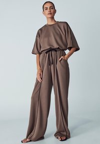 T-shirt marrone oversize con maniche corte e vita regolabile con cordoncino. Pantaloni marroni larghi con tasche laterali e cuciture decorative nere.