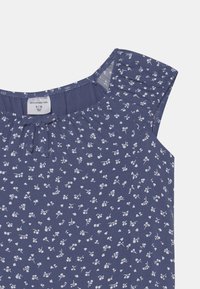 Granatowa bezrękawnik z białym kwiatowym wzorem, zgromadzonym dekoltem i szczegółem w kształcie otworu. Wykonana z miękkiego, lekkiego materiału.