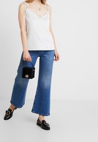 Top bianco senza maniche con spalline regolabili, jeans blu a zampa d'elefante, borsa nera con texture coccodrillo e mocassini neri con fibbia dorata.