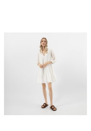Femme debout portant une robe blanche ample à boutons avec des manches trois-quarts et des sandales ouvertes marron, sur un fond blanc uni.