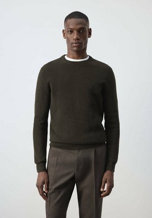 Hombre con suéter de manga larga verde oscuro y pantalones marrones de pie frente a un fondo claro y liso.