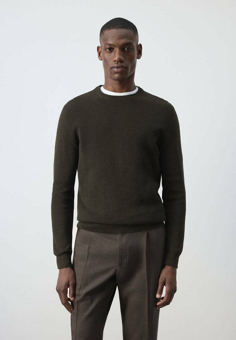 Homme portant un pull à manches longues vert foncé et un pantalon marron, debout devant un fond clair uni.