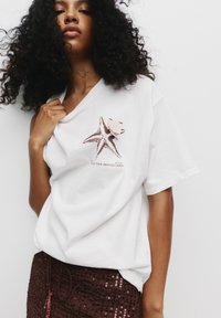 T-shirt en coton blanc à manches courtes avec un motif de starfish marron et du texte, associé à une jupe marron pailletée.