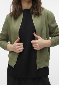 Kvinna med axellångt hår som bär en öppen olivgrön bomberjacka över en svart tröja, håller jackan nära dragkedjan med båda händerna.