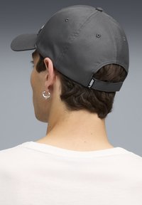Grå baseballkasket med buet skygge, justerbar rem og tekstureret stof. Synlig logo på bagsiden og en sølv hoop ørestik.