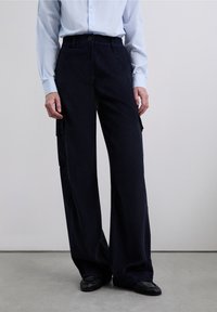 Pantalones anchos de pana en color azul marino con un acabado texturizado, que cuentan con bolsillos laterales y cierre de botón. Combinados con una camisa de color azul claro.