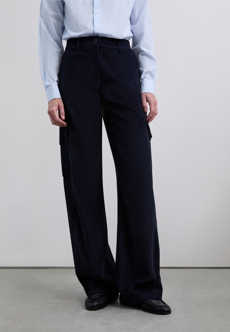Pantalones anchos de pana en color azul marino con un acabado texturizado, que cuentan con bolsillos laterales y cierre de botón. Combinados con una camisa de color azul claro.