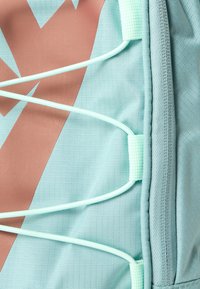 Világoskék szövet texturált felülettel és geometrikus barna mintázattal. Menta zöld elasztikus zsinórok bungee stílusú záródást képeznek az oldalon.