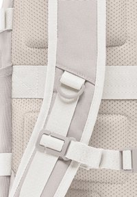 Un sac à dos gris clair avec un panneau en mesh texturé, des bretelles réglables et des accessoires en plastique. Il comprend des sangles d'accentuation blanches et du rembourrage.
