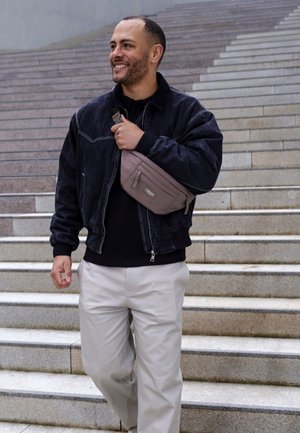 Smiling man wearing black jacket and beige pants carries gray crossbody bag while walking down outdoor stone stairs.