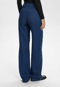 Plati tamsiai mėlynos džinsinės kelnės su aukštu liemeniu. Yra užpakaliniai kišenės su siuvinėjimo akcentais ir lygia tekstūra.