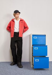 Giacca rossa con zip e dettagli neri sopra una maglietta bianca, pantaloni neri e scarpe nere, accanto a un armadietto blu con due cassetti.