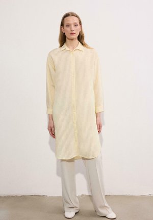 Žena stojaca, oblečená v svetložltých pruhovaných dlho zapínateľných šatách cez krémovo biele široké nohavice a biele topánky, stojaca pred jednoliatou stenou.