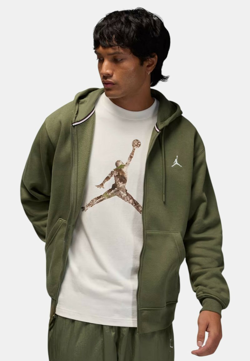 Felpa con cappuccio verde oliva con cerniera, con fodera interna grigia, indossata sopra una t-shirt bianca con una grafica Jumpman in toni e texture naturali.