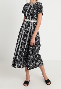 Svart blommig klänning med vita detaljer, korta ärmar och en figursydd midja. Tyget har en slät yta med små blommönster överallt.