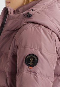 Ljusrosa vadderad jacka med huood, med kviltad struktur och en svart cirkulär logotyp på ärmen med orange accent.