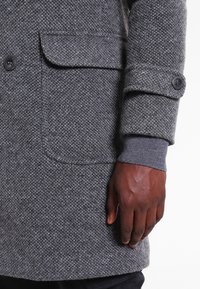 Abrigo texturizado gris con un patrón de espiga, puños abotonados y bolsillos frontales; la tela parece cálida y duradera.