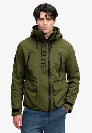 Mand iført grøn hættetrøje med sorte lynlåse og store forlommer, står med hænderne i lommerne på blå jeans.