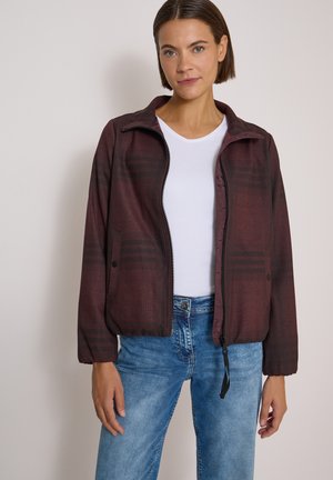 Woman wearing a burgundy zip-up jacket, white t-shirt, and blue jeans, standing against a plain light background.