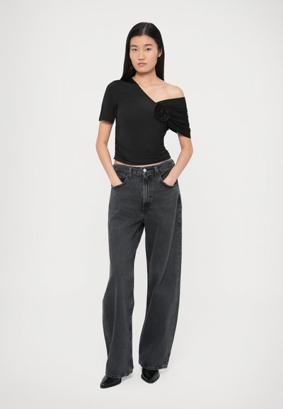 Μαύρο τοπ με ανοιχτούς ώμους και λουλουδάτη λεπτομέρεια στο πλάι, σε συνδυασμό με ψηλόμεσα, άνετα σκούρα γκρι τζιν και μαύρα μυτερά παπούτσια.
