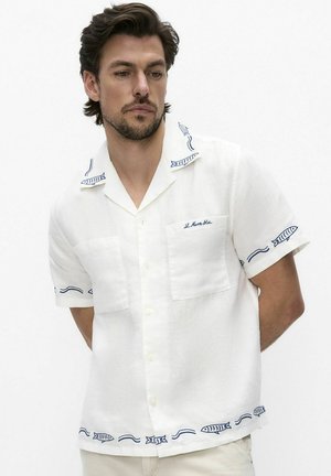 Mies pukeutuneena valkoiseen, lyhythihaiseen napitettuun paitaan, jossa on sinisiä kirjailtuja kaloja ja aaltoilevia viivakuvioita kauluksessa, hihoissa ja helmassa.