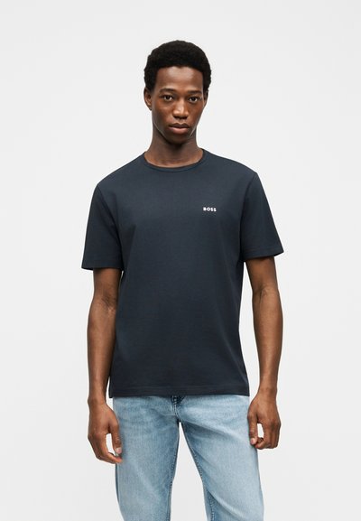 Homme portant un t-shirt noir à manches courtes avec le logo « BOSS » et un jean bleu clair, debout devant un fond blanc uni.