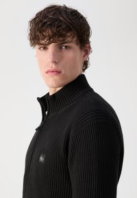 Maglione nero con zip, in tessuto a coste e colletto alto. Presenta un piccolo logo sul petto, realizzato in un tessuto morbido.