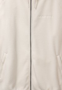 Kremowa bluza polarowa zapinana na zamek, z bocznymi kieszeniami i haftowanym napisem „EST SCANDINAVIA” na lewej piersi.