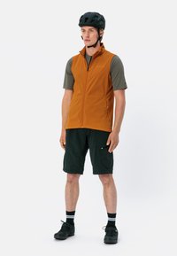 Oranges ärmelloses Oberteil über einem grauen T-Shirt mit kurzen Ärmeln, kombiniert mit schwarzen Shorts, schwarzen Socken und Radschuhen; Modell trägt einen schwarzen Helm.