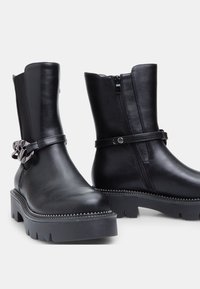 Botas de tobillo de cuero negro con suela gruesa, cremallera lateral y una correa decorativa con un acento metálico. Textura suave y diseño resistente.