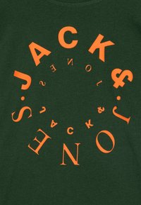 T-shirt en coton vert avec des lettres orange audacieuses disposées en motif circulaire, affichant du texte et des symboles, avec une texture douce.
