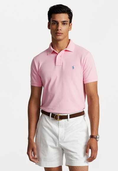 Camisa polo de color rosa claro con mangas cortas y cuello, que presenta un logo azul. Combinada con pantalones cortos blancos y un cinturón marrón.