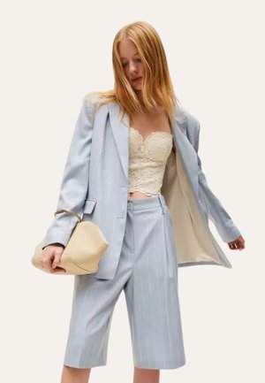 Donna con blazer azzurro chiaro a righe sottili e pantaloncini lunghi abbinate, top corto in pizzo crema, che tiene una borsa beige con manico dorato.