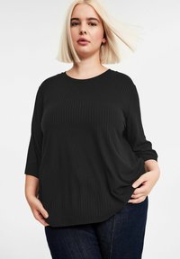 Blonde Frau mit glattem Haar, die ein schwarzes, geripptes Oberteil mit dreiviertel Ärmeln und dunklen blauen Jeans trägt und mit den Händen in den Taschen posiert.