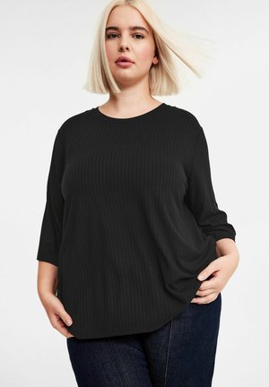 Blond kvinde med glat hår iført en sort ribbet bluse med trekvartærmer og mørkeblå jeans, poserende med hænderne i lommerne.