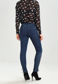 Modré skinny džíny s vysokým pasem, hladkým povrchem a šněrováním podél dolních nohavic, spárované s průhlednou černou halenkou s hvězdicovým vzorem.