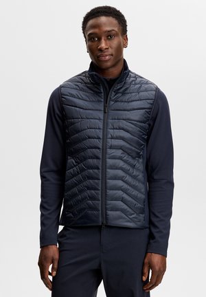 Man draagt een marineblauw doorgestikt vest over een marineblauw shirt met lange mouwen, staand tegen een effen lichtgrijze achtergrond.