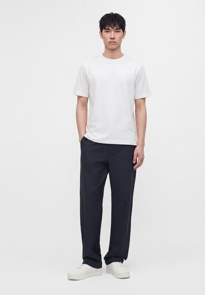 Jeune homme debout, les mains dans les poches, portant un t-shirt blanc à manches courtes, un pantalon foncé et des baskets blanches sur un fond uni.