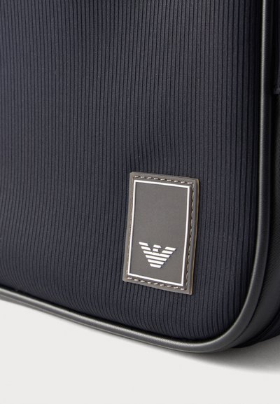 Κοντινή λήψη σκοτεινής υφασμάτινης τσάντας με ριμπ υφή και ραμμένη ορθογώνια ετικέτα λογότυπου, που διαθέτει σχεδιασμό με στυλιζαρισμένο αετό στο κέντρο στο κάτω μέρος.