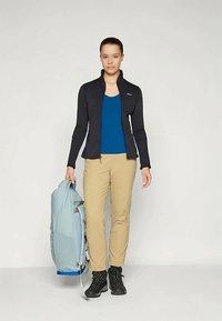 Giacca nera con zip, top blu, pantaloni beige, stivali da trekking neri. Tieni uno zaino azzurro chiaro. Abbigliamento outdoor semplice e funzionale.