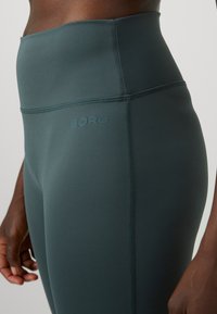 Visoko pasovi, teal leggingi izdelani iz gladkega, elastičnega materiala. Ponuja šivano šiv in embosirano oznako "BORG" na strani.