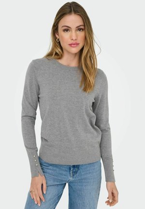 Pull en maille gris à manches longues, décolleté rond et boutons décoratifs aux poignets. Associé à un jean en denim bleu.