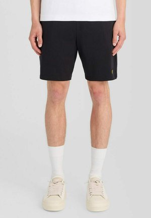 Mann trägt schwarze Shorts, weiße Socken und beige Turnschuhe und steht vor einem schlichten hellen Hintergrund.