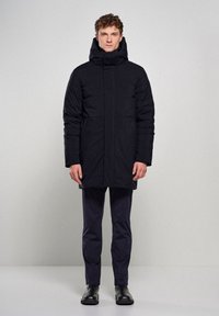 Sort polstret frakke med høj krave og hætte, udstyret med to frontlommer, slankt design og vandafvisende materiale. Slim-fit bukser og støvler.