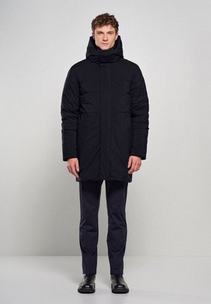 Sort polstret frakke med høj krave og hætte, udstyret med to frontlommer, slankt design og vandafvisende materiale. Slim-fit bukser og støvler.