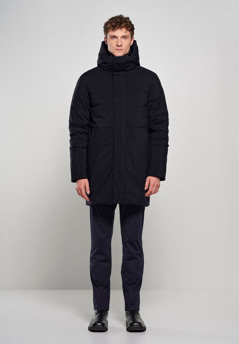 Sort polstret frakke med høj krave og hætte, udstyret med to frontlommer, slankt design og vandafvisende materiale. Slim-fit bukser og støvler.