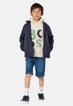 Marineblaue Jacke mit strukturierter Oberfläche, beigefarbene Kapuzenjacke mit farbigem Motiv, Jeansshorts, weiße Socken und marineblaue Sneakers. Einfaches, lässiges Outfit.